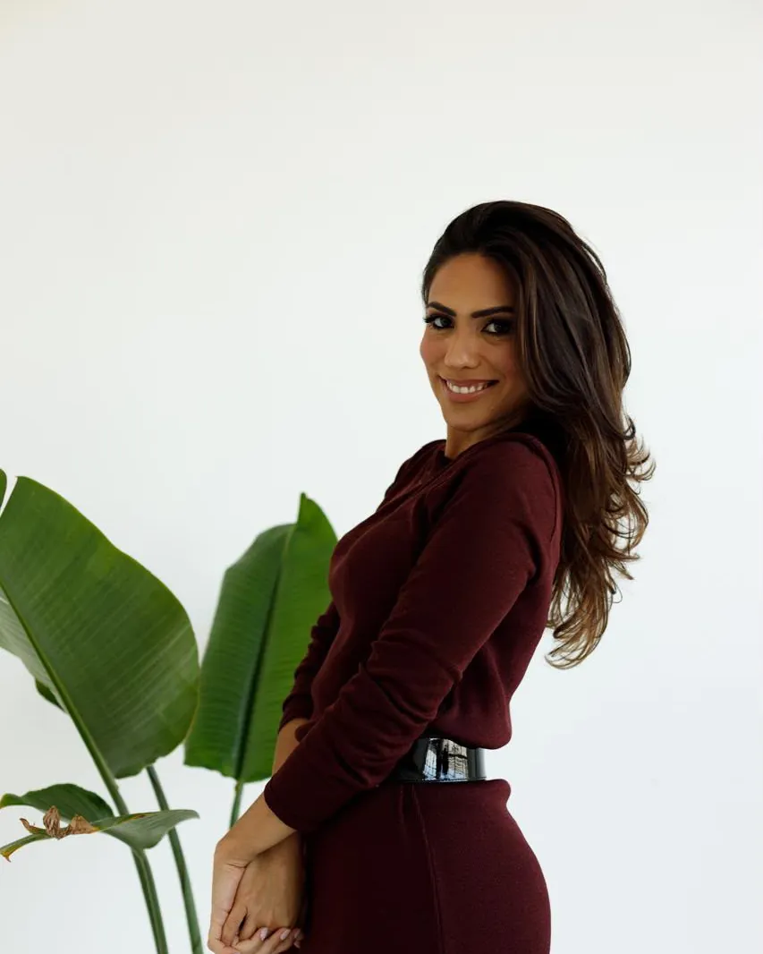 Dra. Dieme Schettert sorrindo, de blusa vinho, ao lado de uma planta